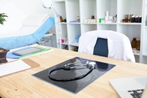 Stethoscope on a desk in a medical office, with a white coat hanging on a chair and medical supplies visible in the background, illustrating health and wellness services.